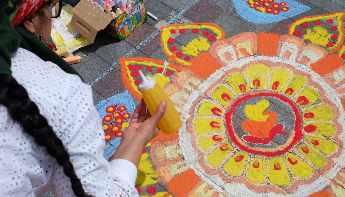 Participants sit on the pavement next to colorful Rangoli designs made with chalk, showcasing intricate patterns and symbols.