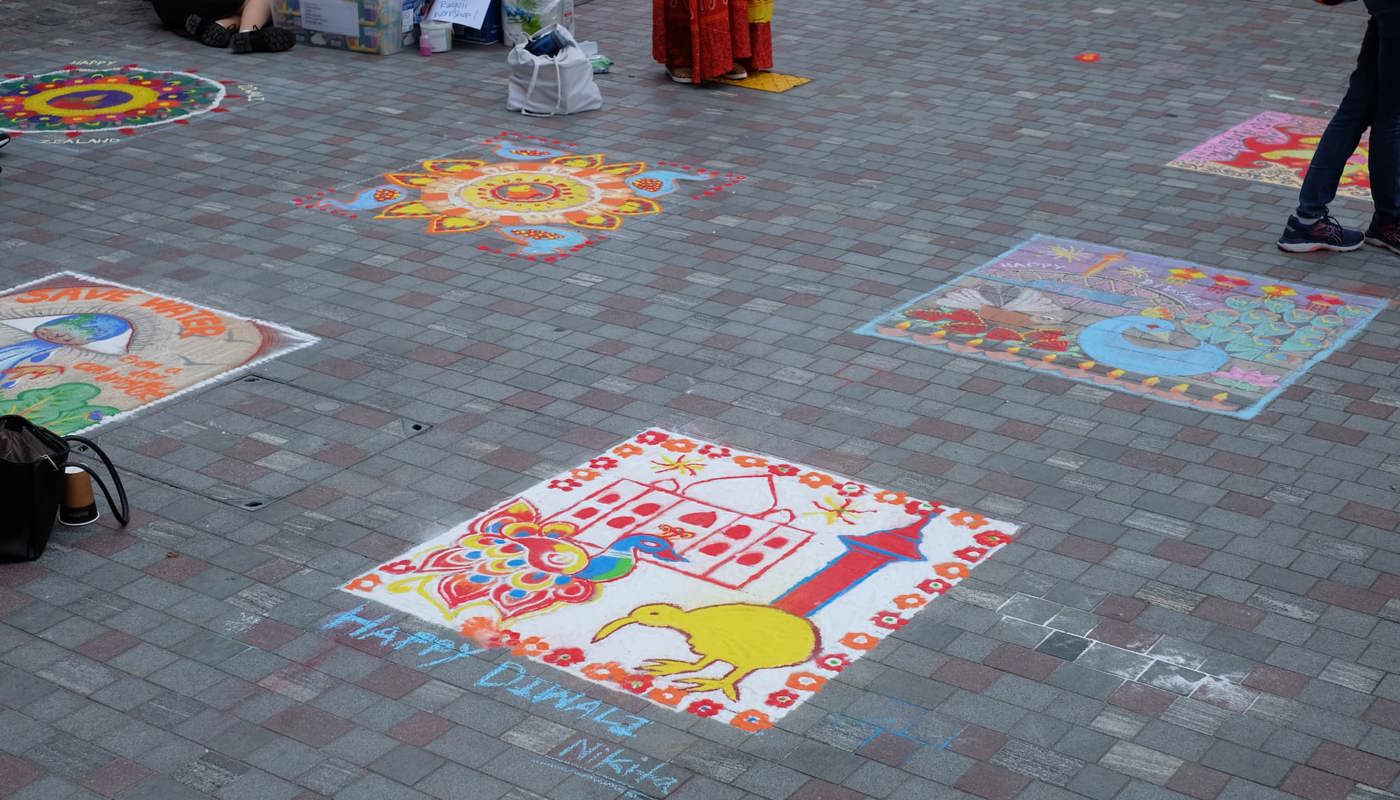 Several chalk Rangoli designs are displayed on a brick-paved area, featuring colorful patterns and cultural symbols, such as a yellow kiwi bird