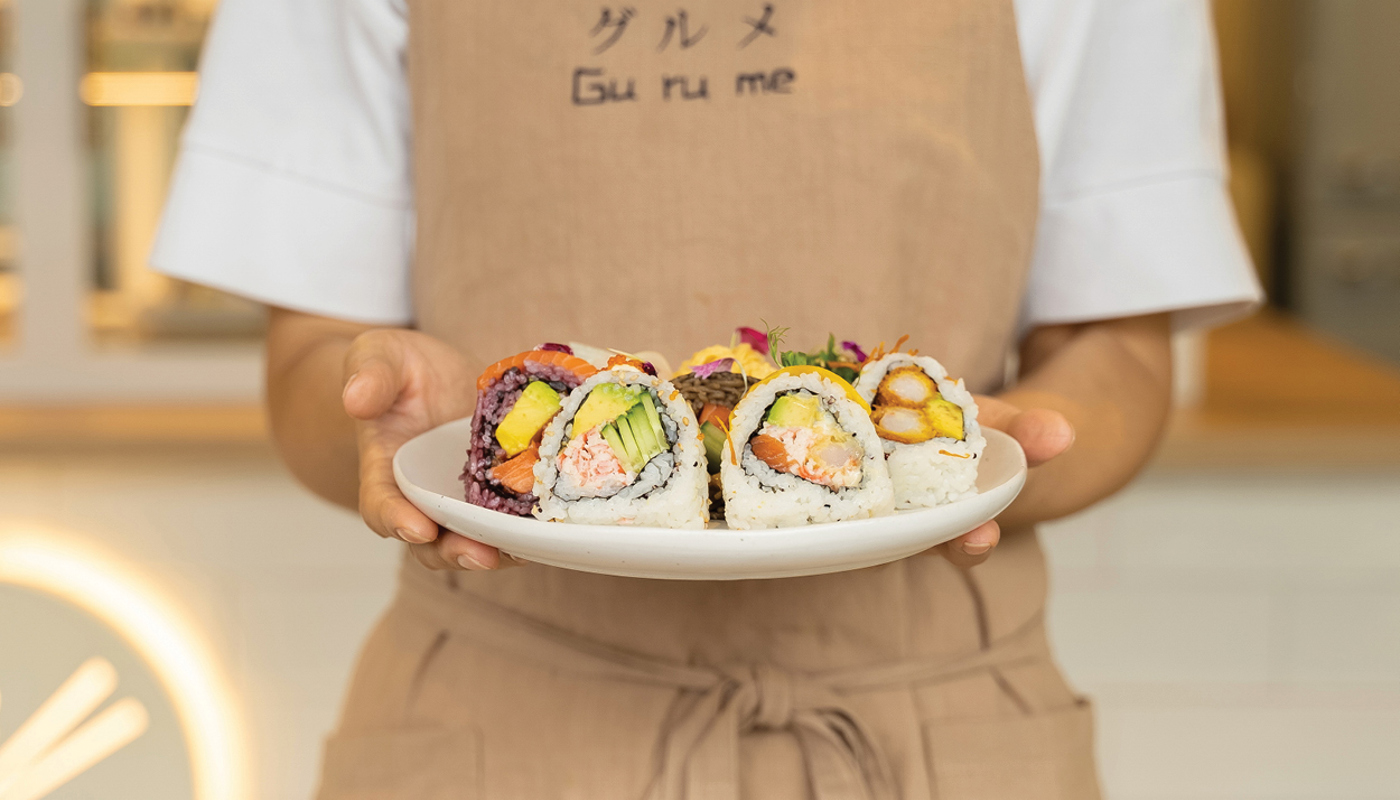 A person holds a plate of sushi whilst wearing a Gurume branded apron.
